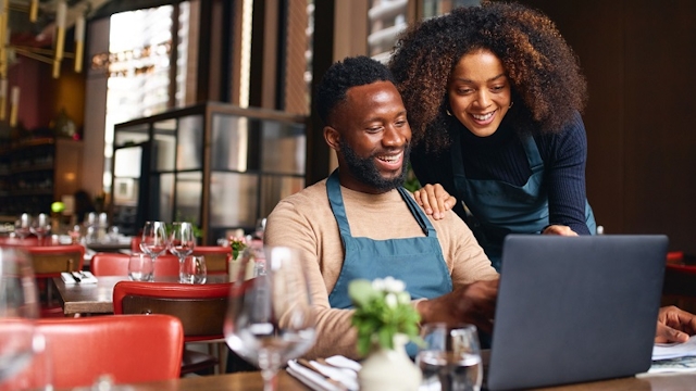 Two happy Black male and female restaurant owners sit at a table and look at a laptop