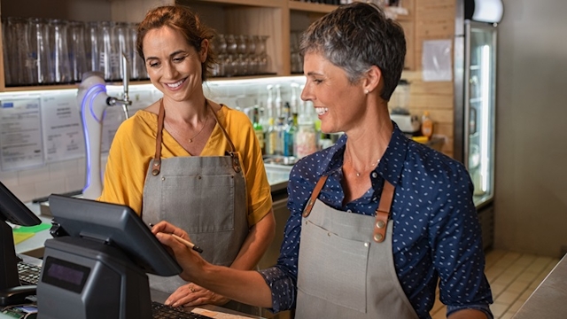 A mature female coffee shop owner showing a new female employee how to use the till