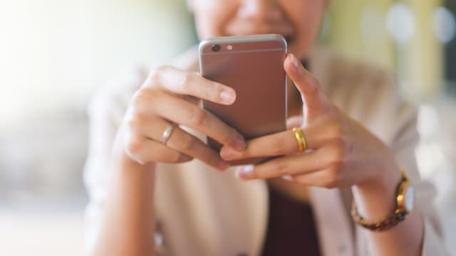Person holding a smartphone with both hands, focusing on the screen. They are wearing rings and a beige jacket.