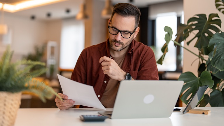 A bearded male business owner wearing glasses and a dark red shirt working in a plant-filled office, laptop open and looking at a document