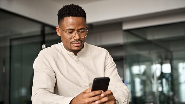 Black man in glasses and a beige shirt looking at a smartphone, sitting in a modern office setting.