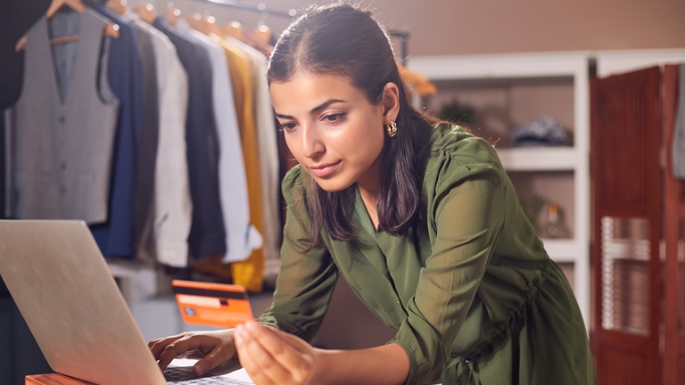 A young female Indian/Asian fashion business owner holds a credit card while she makes an online payment on her laptop