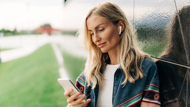 Woman with blonde hair wearing earbuds, looking at her smartphone while leaning against a shiny wall outdoors.
