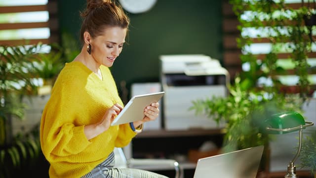 Woman in a yellow sweater uses a tablet in a bright office with plants and a printer in the background.
