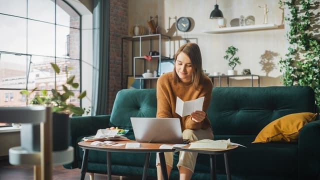 Woman sits on a green sofa, reading a paper, with a laptop on a wooden table in a cozy, modern living room.
